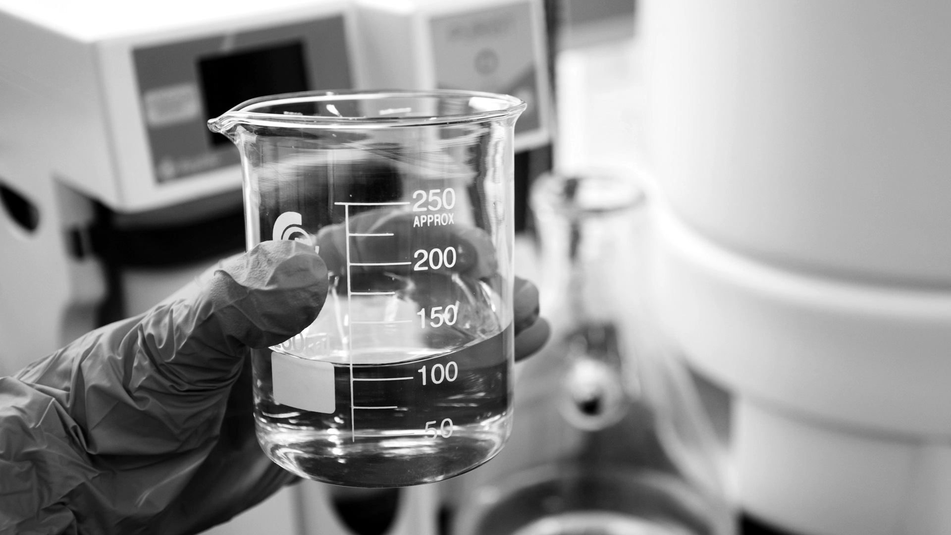A gloved hand holding a glass beaker filled with clear liquid in a laboratory.