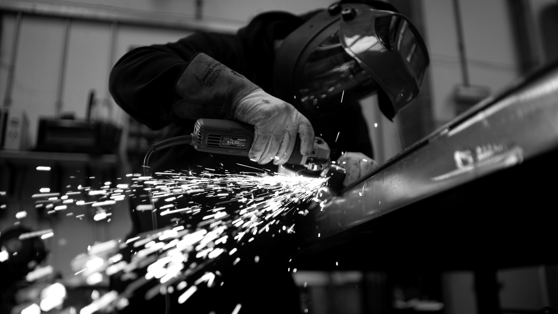 A person wearing a welding helmet grinding a metal beam with sparks flying.