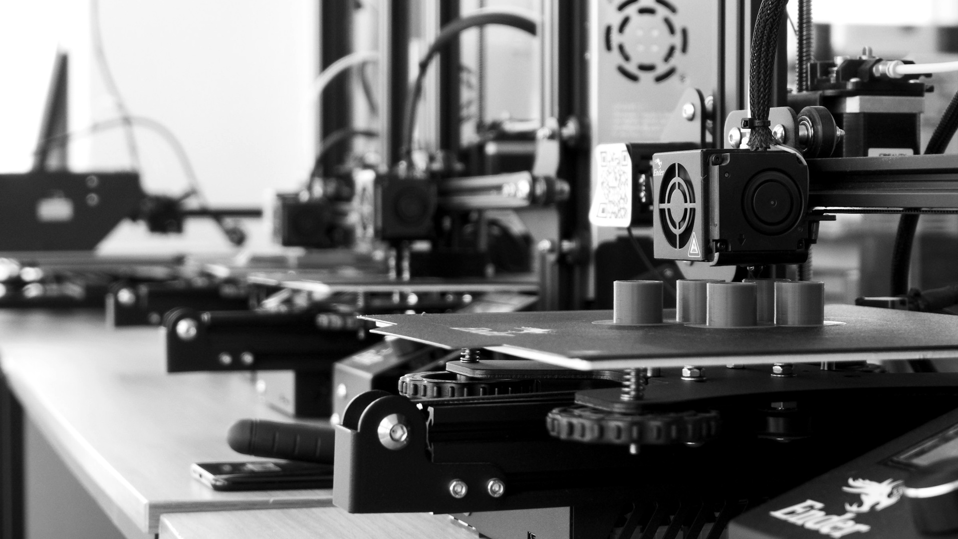 A row of 3D printers creating blue plastic cylinders on a wooden workbench.