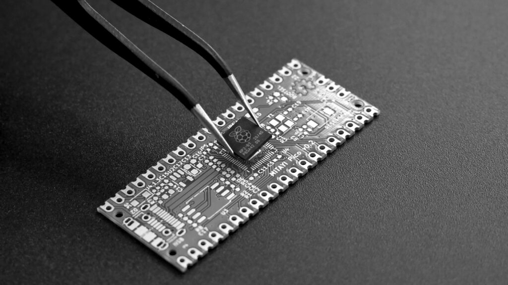 Tweezers placing a small black microchip onto a green circuit board.