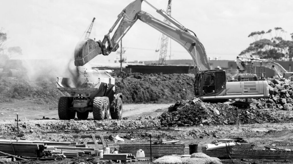 A teal excavator dumping soil into a yellow dump truck at a construction site.